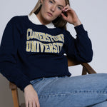 Person wearing a navy blue crewneck with 'Cornerstone University' text, sitting on a wooden chair.
