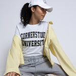 Woman wearing a 'Cornerstone University' t-shirt with a white cap and light yellow jacket on a plain background