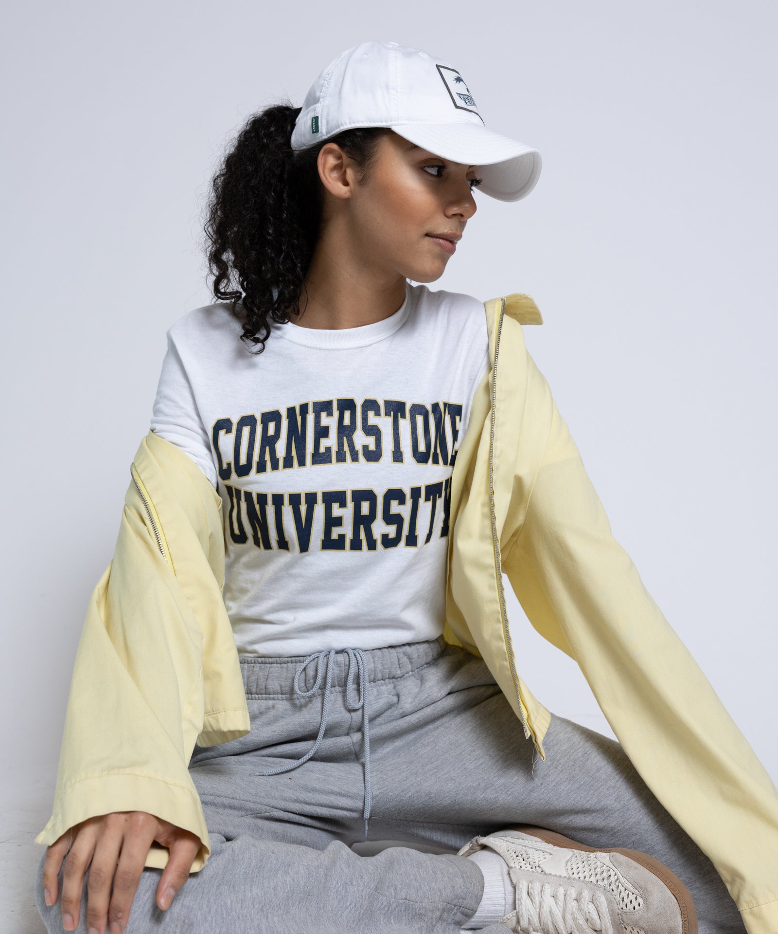 Woman wearing a 'Cornerstone University' t-shirt with a white cap and light yellow jacket on a plain background