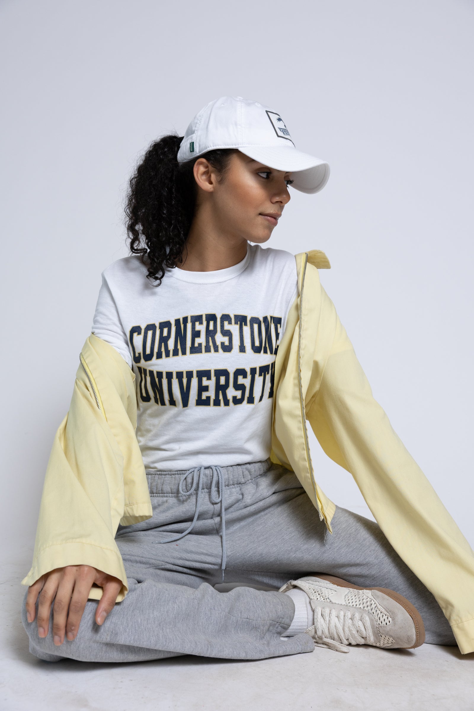 Woman wearing a 'Cornerstone University' t-shirt with a white cap and light yellow jacket on a plain background