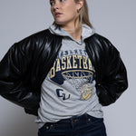 Person wearing a black leather jacket over a gray hoodie with Cornerstone basketball graphics on a plain background