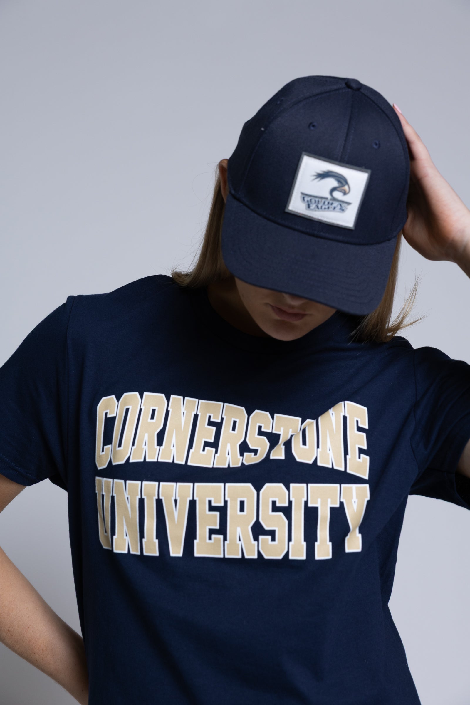 Woman wearing a navy blue t-shirt with 'Cornerstone University' text and a navy hat with a plain background behind her